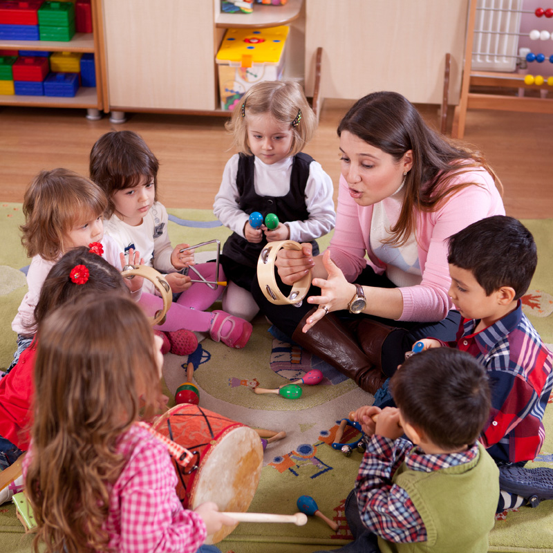 Childcare teacher sitting on floor with a circle of children. They are all exploring handheld musical instruments, inlcuding tamborines, shakers, and drums.
